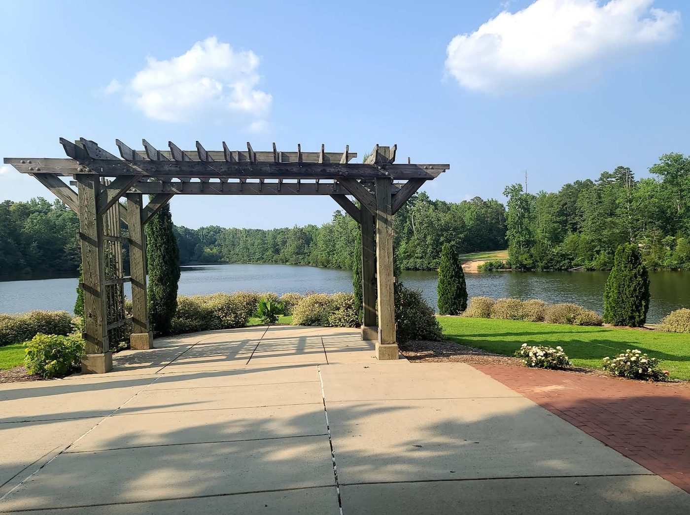 Wooden pergola on a patio beside a lake, with trees and blue sky